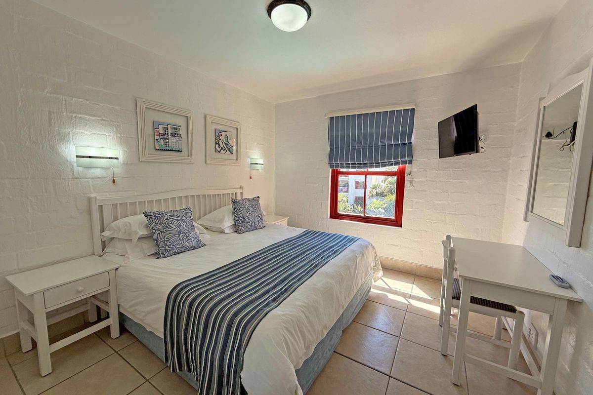 A bright bedroom featuring white painted brick walls, tiled flooring, a window with a red frame, and a ceiling-mounted light fixture.