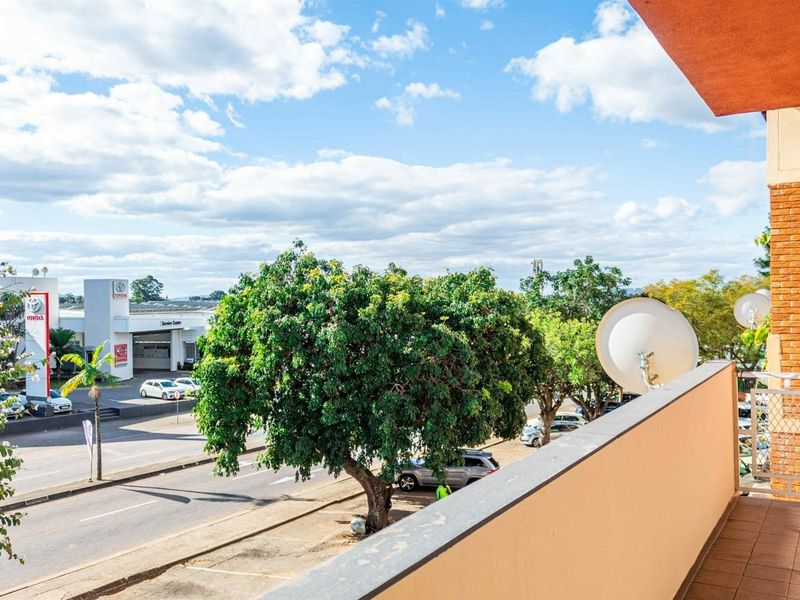 A view from an apartment balcony overlooking a street and commercial area, featuring a tiled floor and solid perimeter wall.