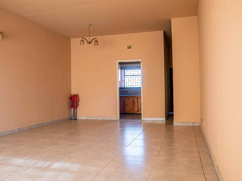 A spacious, open-plan living area featuring neutral tiled flooring and a central light fixture, with a direct view into the adjacent kitchen.