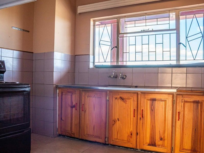 A dated kitchen featuring wooden cabinetry, tiled splashbacks, a stainless steel sink, and a freestanding stove, with security bars on the window.