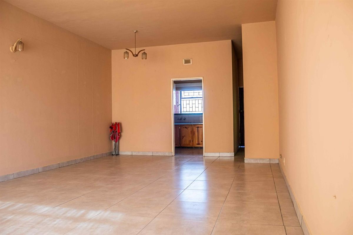 A spacious, open-plan living area featuring neutral tiled flooring and a central light fixture, with a direct view into the adjacent kitchen.