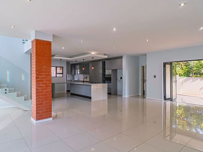 Spacious open-plan living area featuring a modern kitchen with island, exposed brick pillar, glass staircase, and bi-fold doors leading to the outdoor space.