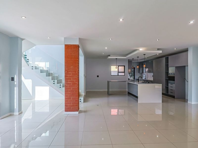 Spacious open-plan living area featuring a modern kitchen with island, sleek cabinetry, and a striking exposed brick pillar, complemented by a contemporary glass-railed staircase.