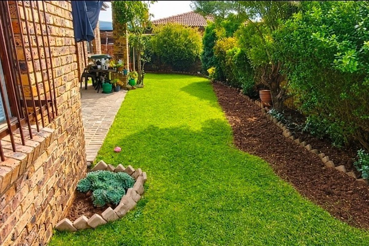 A well-maintained rear garden featuring a paved patio area, lush green lawn, and established garden beds with brick edging.