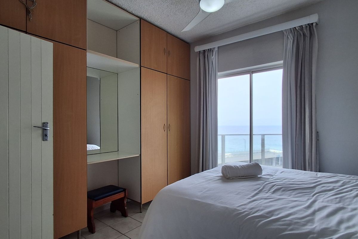 A bedroom featuring built-in wooden wardrobes with an integrated vanity area, tiled flooring, and sliding glass doors opening to a balcony with ocean views.