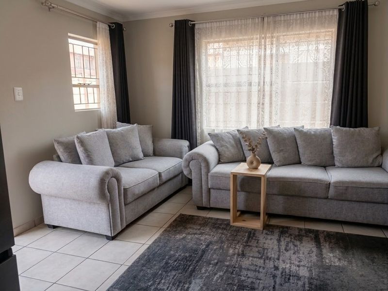 A comfortable living room featuring neutral tiled flooring, large windows with security bars, and modern curtain fixtures, creating a bright and inviting space.