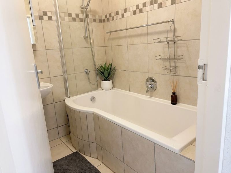 A clean and functional bathroom featuring a tiled bathtub with an integrated shower mixer, glass screen, and neutral wall and floor tiling.