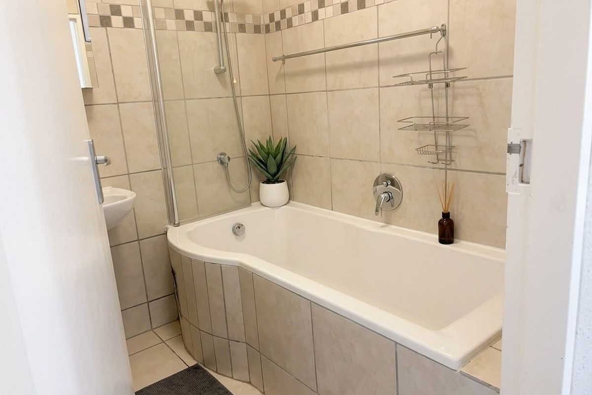 A clean and functional bathroom featuring a tiled bathtub with an integrated shower mixer, glass screen, and neutral wall and floor tiling.