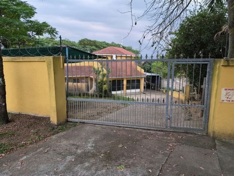A gated residential property entrance featuring a concrete driveway, perimeter wall with security fencing, and a view of the main house structure.