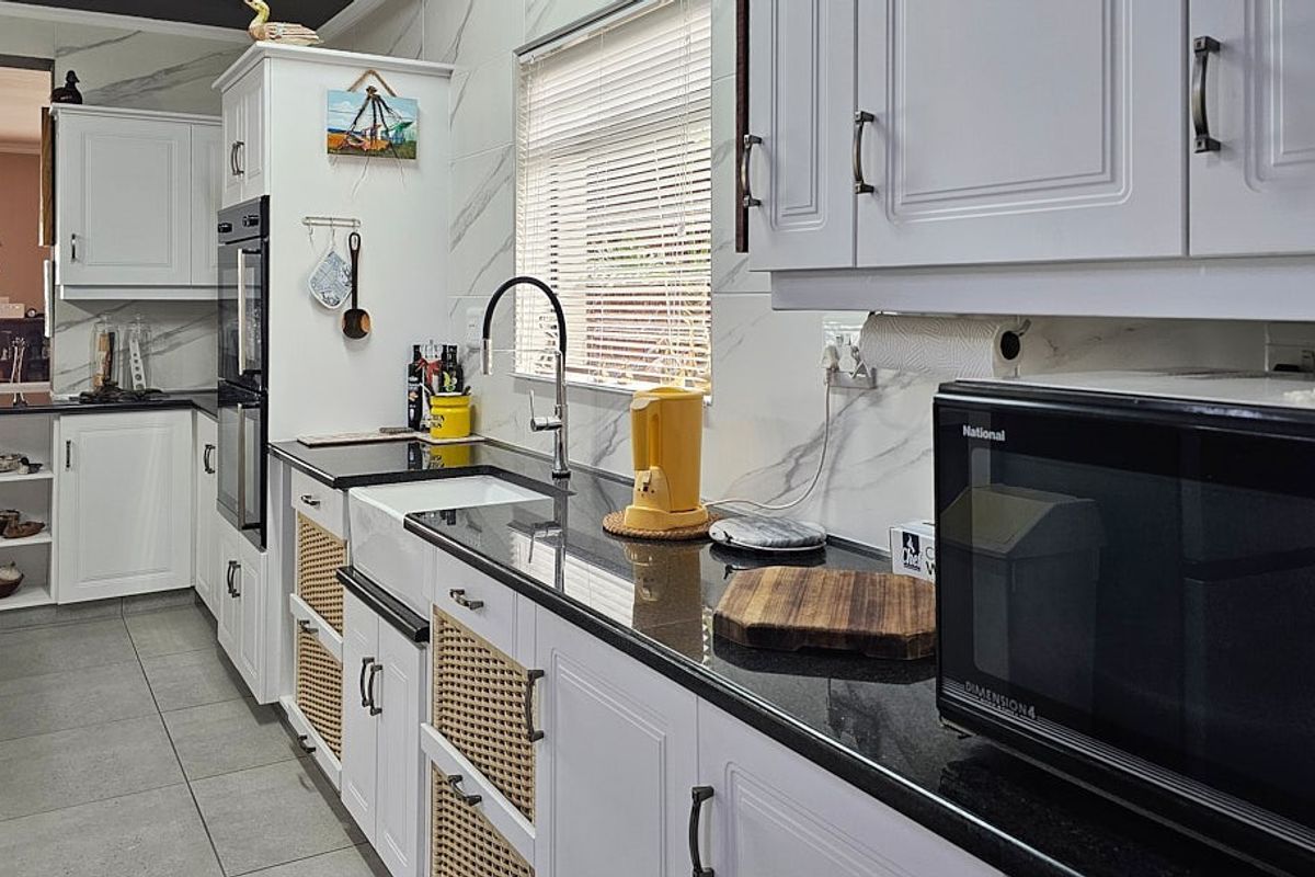 Modern kitchen featuring white cabinetry with rattan accents, dark granite countertops, a farmhouse-style sink, and marble-look wall tiling.