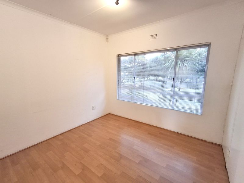 A bright, empty office space featuring laminate flooring, neutral walls, and large windows fitted with venetian blinds.