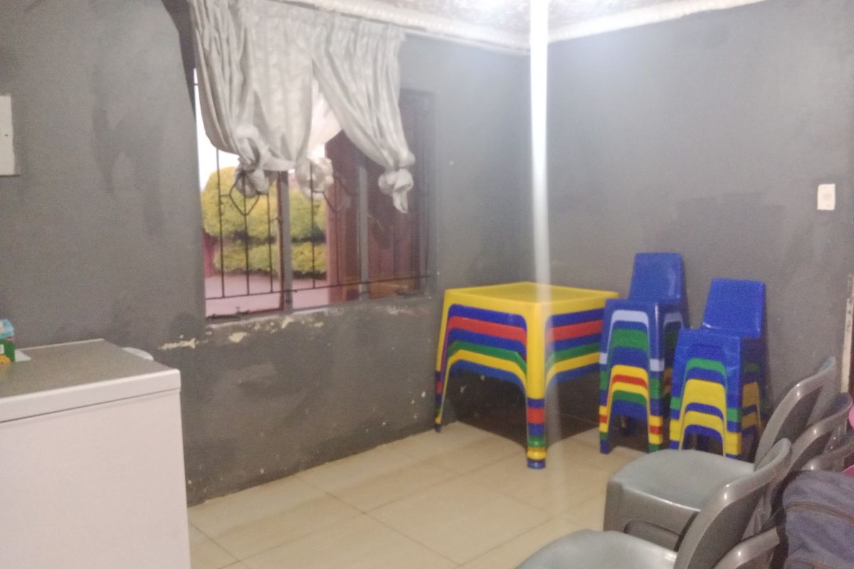 A simple interior living space featuring tiled flooring, a window with security bars, and plain grey walls in need of maintenance.