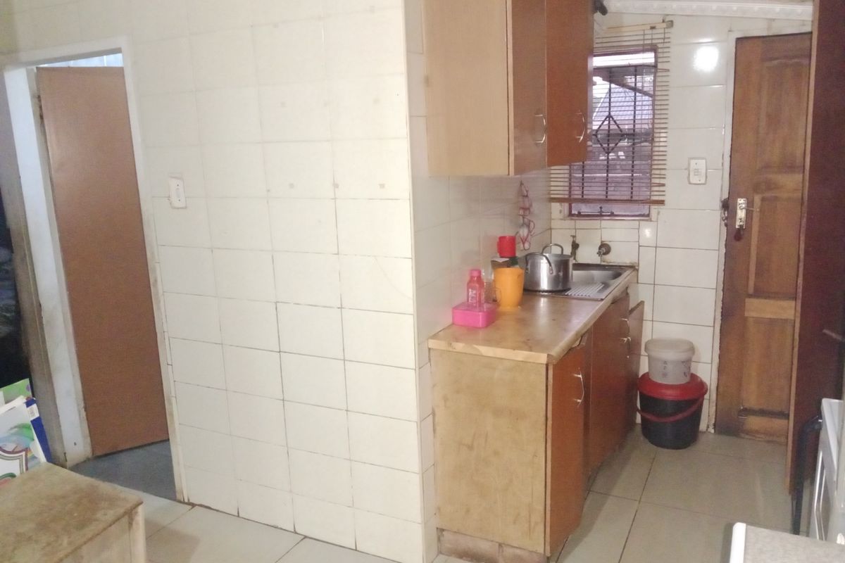 A compact kitchen area featuring tiled walls, wooden cabinetry, and a stainless steel sink, requiring cosmetic updates to modernize the space.