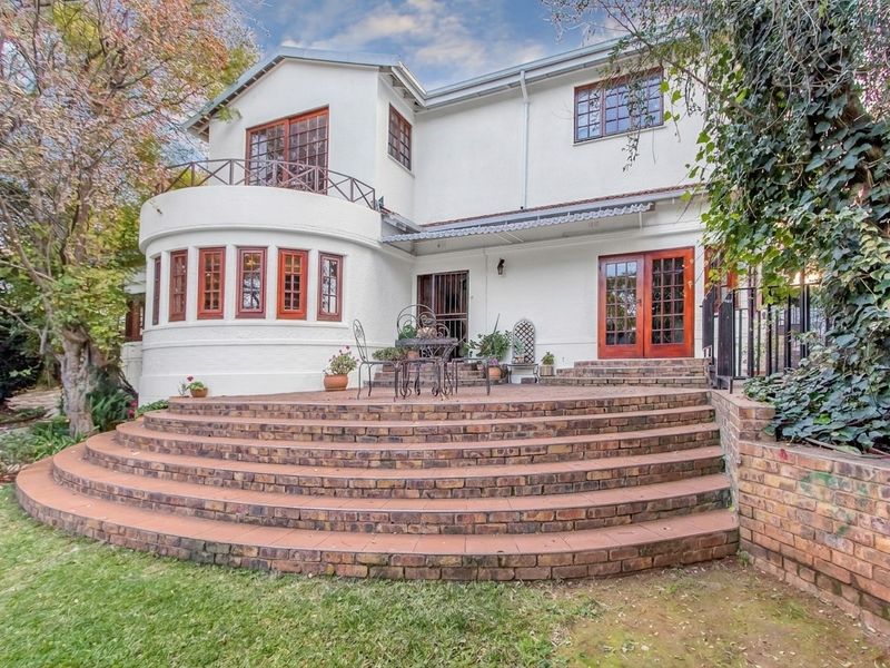 Rear elevation of a two-story house featuring a large curved brick patio, wooden window frames, and a balcony overlooking the garden.
