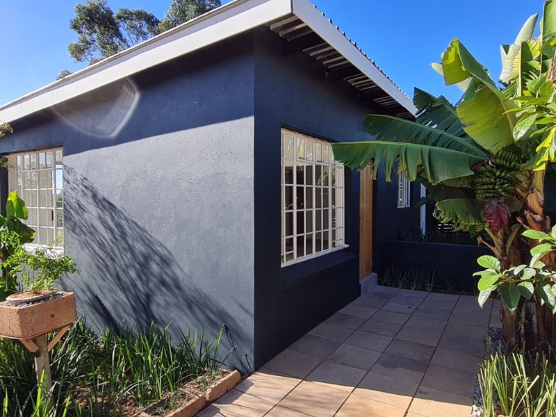 A well-maintained cottage exterior featuring dark painted walls, grid-pattern windows, and a paved patio area surrounded by lush garden landscaping.