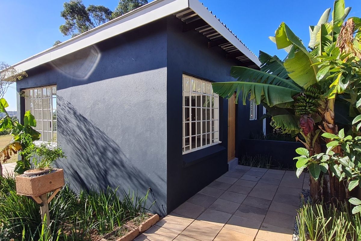 A well-maintained cottage exterior featuring dark painted walls, grid-pattern windows, and a paved patio area surrounded by lush garden landscaping.