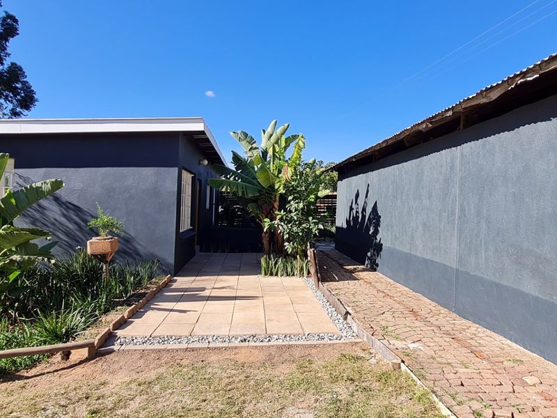 A rear exterior view of the cottage featuring a tiled patio area, brick pathway, and landscaped garden beds against dark painted walls.