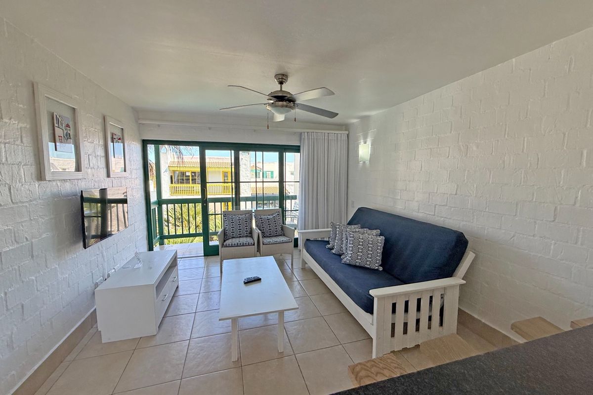 A bright living room featuring white painted brick walls, tiled flooring, a ceiling fan, and sliding glass doors leading to a balcony.