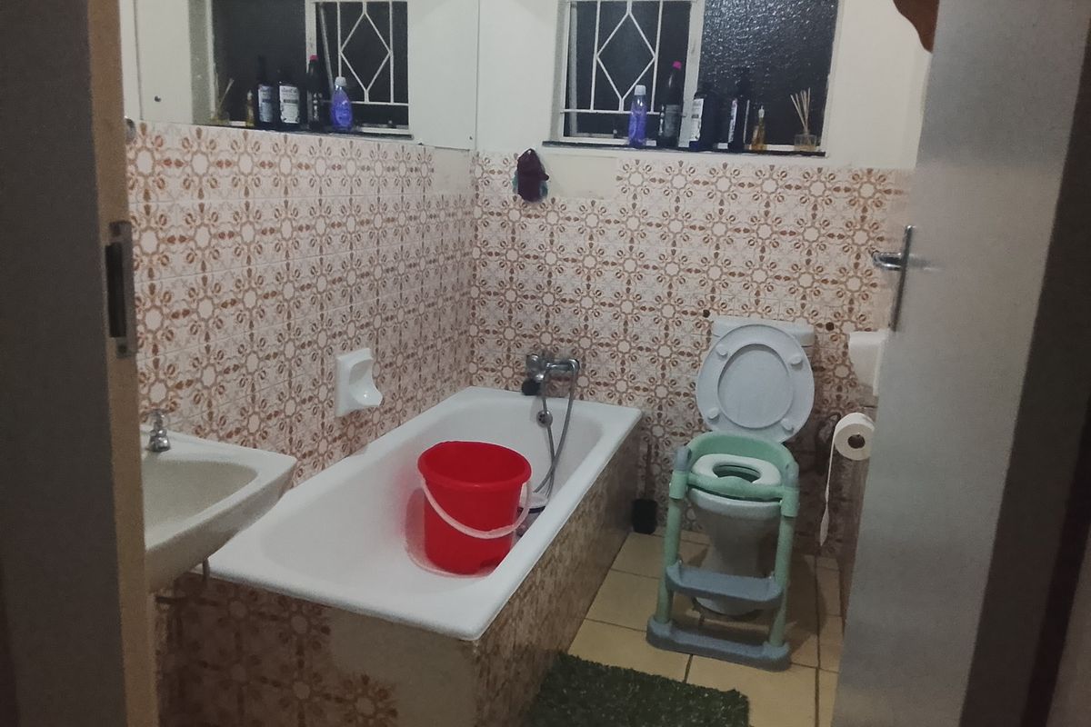 A dated bathroom featuring a standard bathtub, toilet, and washbasin with patterned wall tiles and security bars on the windows.