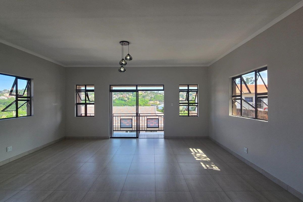 A spacious, empty living area featuring neutral tiled flooring, modern pendant lighting, and large windows providing ample natural light and scenic views.