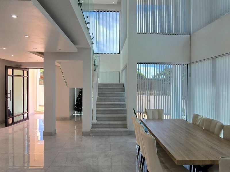 Spacious open-plan living and dining area featuring high ceilings, large windows with vertical blinds, tiled flooring, and a modern staircase with glass balustrades.