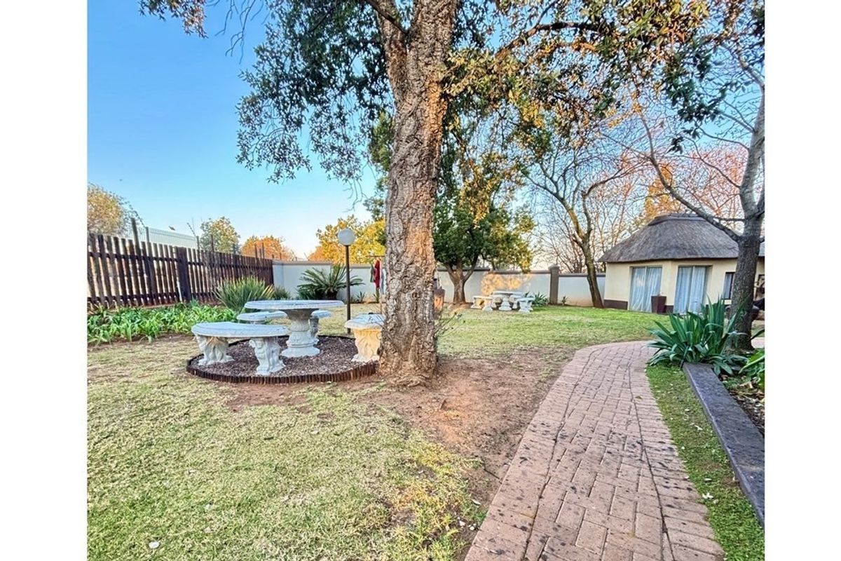 A spacious garden area featuring a paved walkway, mature trees, and a small thatched-roof structure in the background.