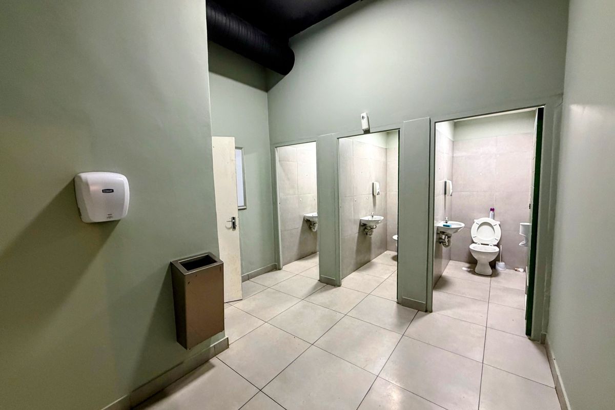 Communal office bathroom featuring multiple toilet cubicles, wall-mounted basins, tiled flooring, and an industrial-style exposed ceiling.