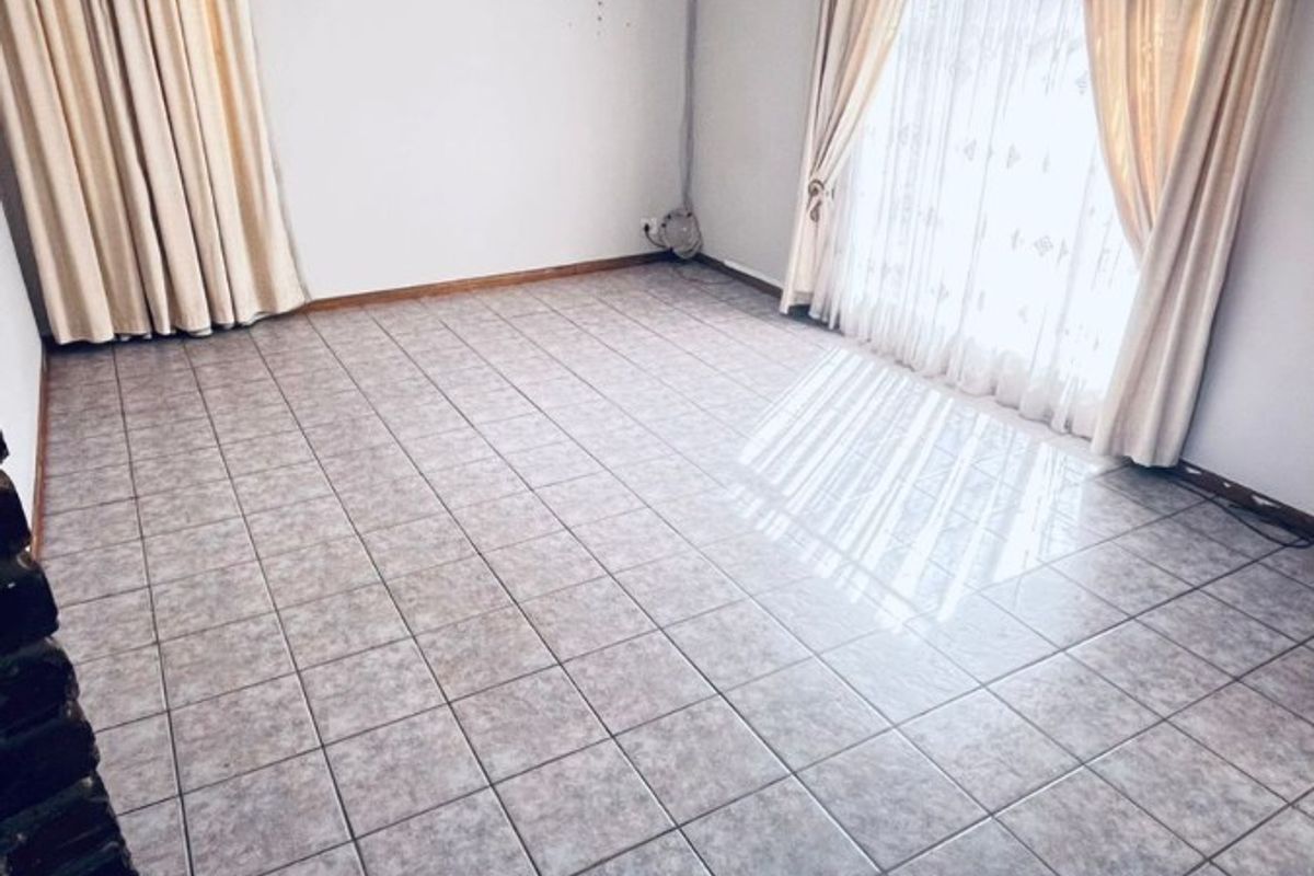 A bright, empty living room featuring neutral ceramic floor tiles, a classic pendant light fixture, and large windows dressed with floor-to-ceiling curtains.