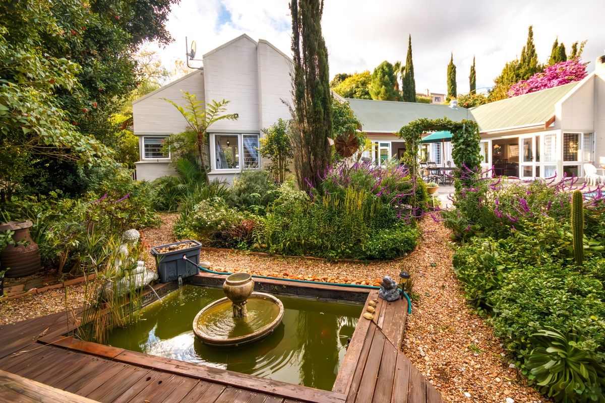 A lush rear garden featuring a central water fountain, gravel pathways, established landscaping, and a wooden deck area adjacent to the home's rear facade.