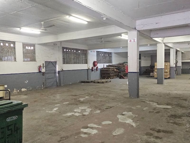 Spacious industrial warehouse area featuring concrete flooring, exposed structural columns, fluorescent lighting, and a secure metal mesh enclosure.
