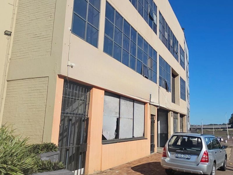 Exterior view of a multi-story industrial building featuring large steel-framed windows, security gates, and a paved frontage area.