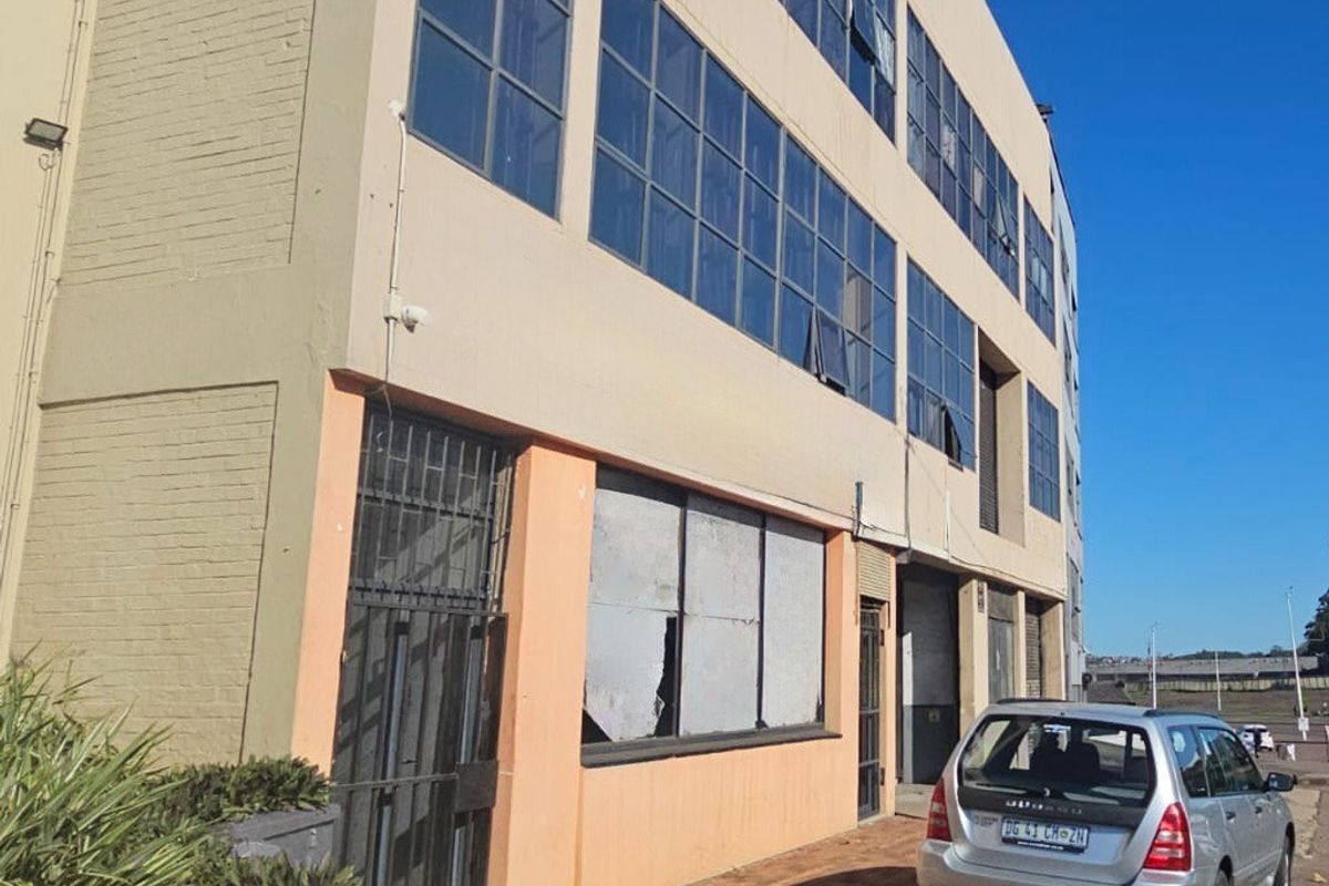 Exterior view of a multi-story industrial building featuring large steel-framed windows, security gates, and a paved frontage area.