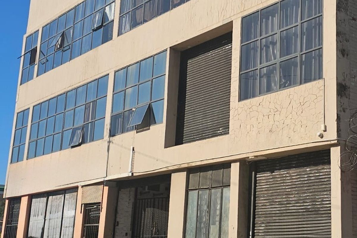Multi-story industrial building facade featuring large grid-style windows, roller shutter doors, and secure metal entrance gates.