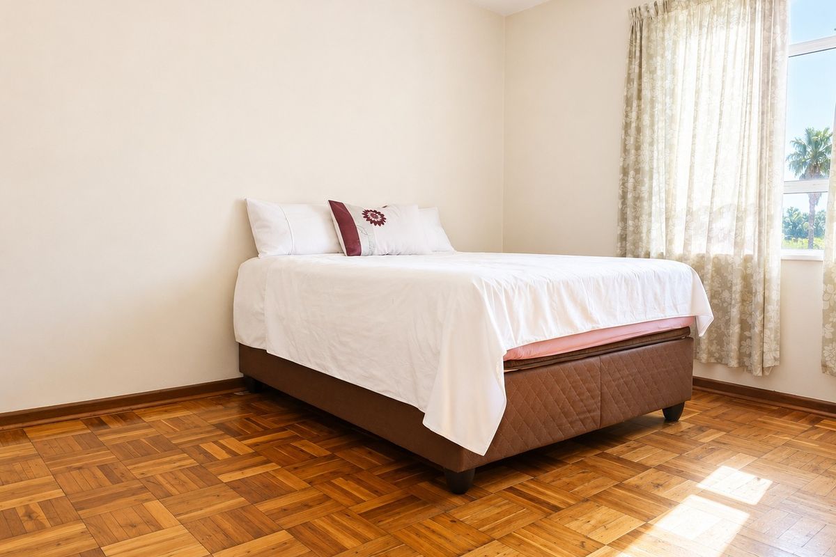 A bright bedroom featuring warm-toned parquet flooring and a large window that allows for ample natural light.