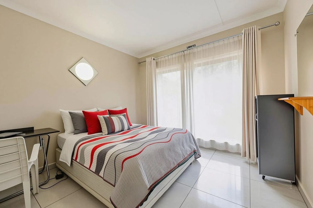A bright and well-maintained bedroom featuring neutral wall tones, large sliding glass doors with curtains, and polished floor tiles.