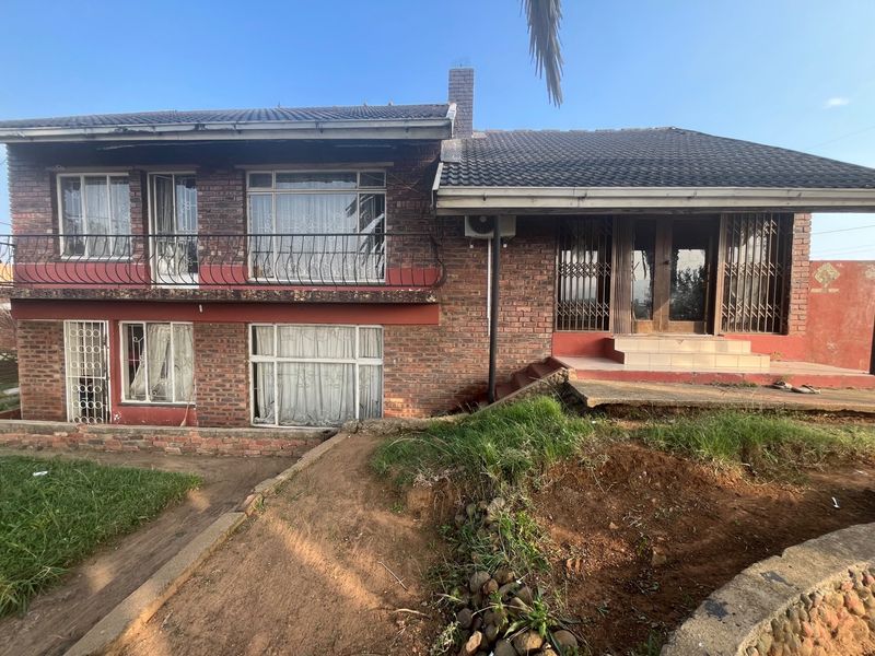 A two-story face-brick house featuring a balcony, tiled roof, and security gates, set on a sloped plot requiring landscaping and exterior maintenance.