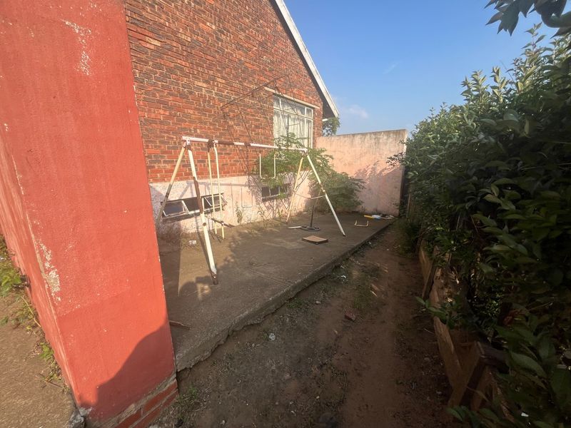 A rear exterior view of the property featuring a concrete patio area, brick facade, and surrounding garden space in need of maintenance.