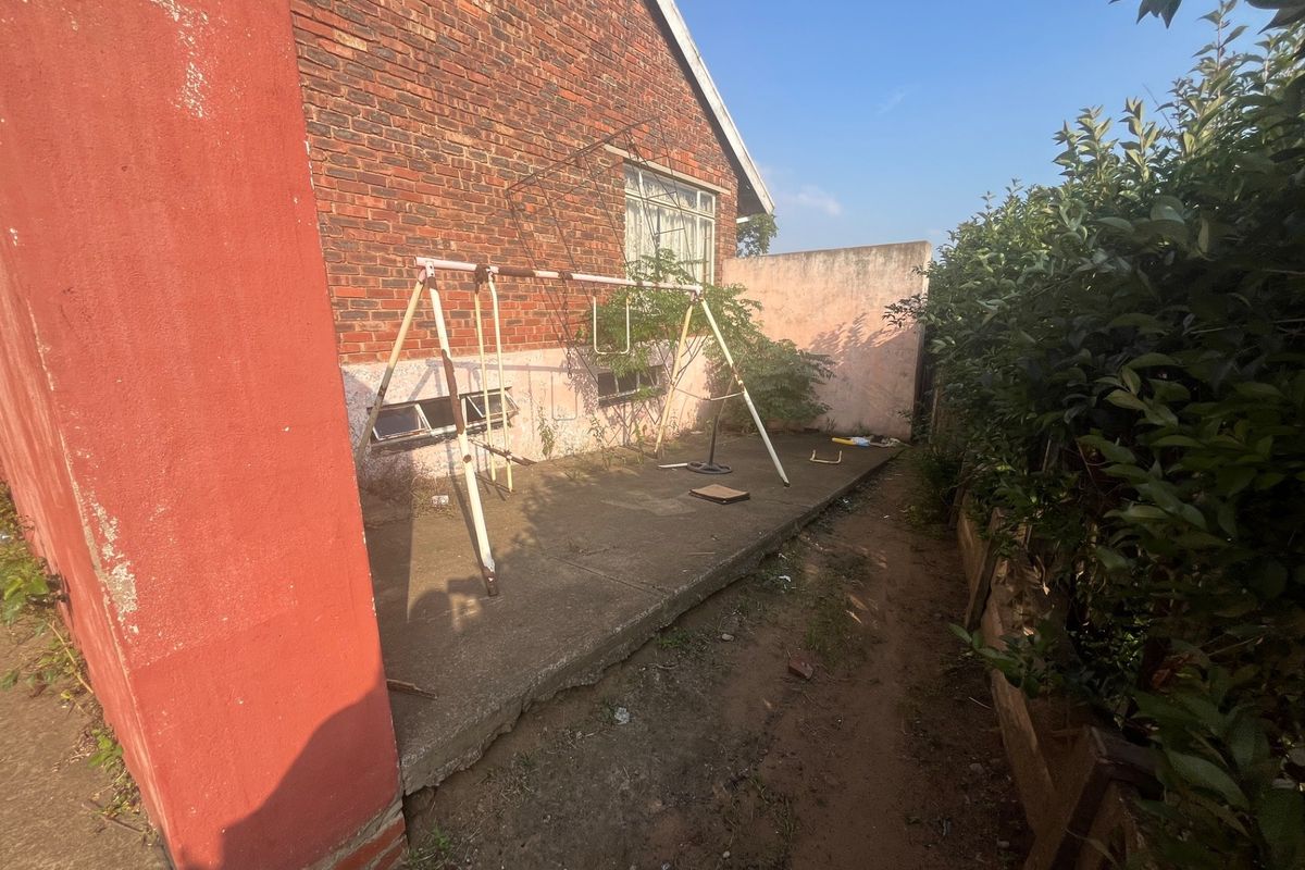A rear exterior view of the property featuring a concrete patio area, brick facade, and surrounding garden space in need of maintenance.