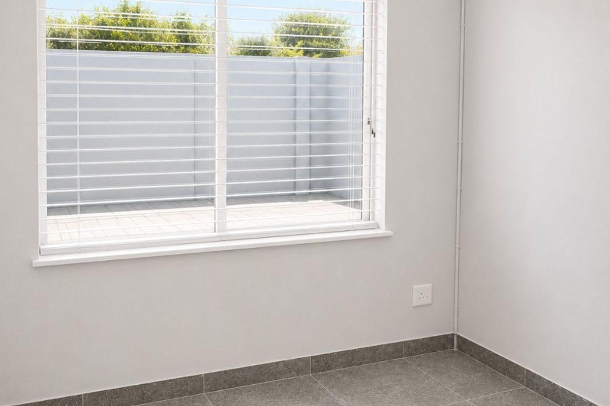 A clean, empty bedroom featuring neutral grey tiled flooring, white walls, and a large window fitted with venetian blinds.