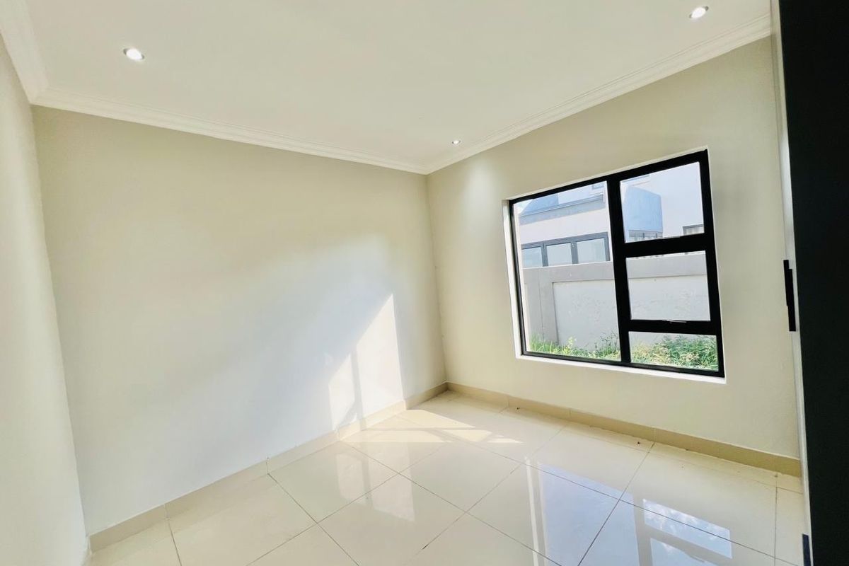 A bright, empty bedroom featuring neutral tiled flooring, recessed ceiling lighting, and a large window with a black frame.