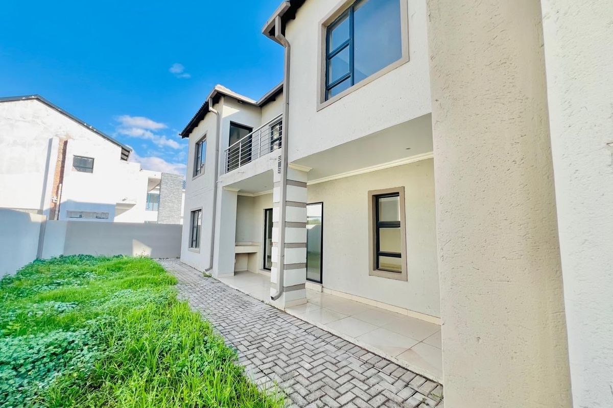 Rear view of a two-story residential property featuring a paved patio area, built-in braai, and a balcony with metal railings.