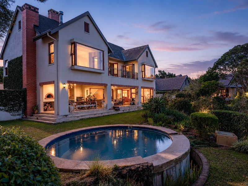 Stunning rear elevation of a multi-story home featuring a circular swimming pool, covered patio with built-in pizza oven, and a beautifully landscaped garden at dusk.