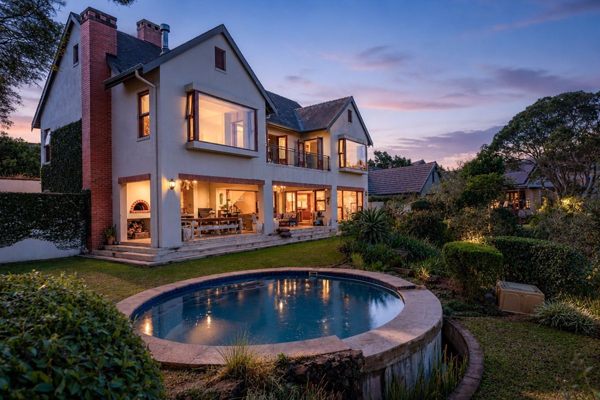 Stunning rear elevation of a multi-story home featuring a circular swimming pool, covered patio with built-in pizza oven, and a beautifully landscaped garden at dusk.