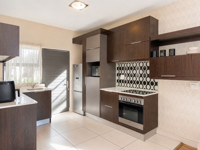 Modern kitchen featuring dark wood cabinetry, integrated oven and hob, patterned tile backsplash, and light-colored floor tiles with direct exterior access.