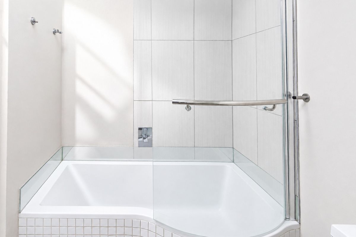 A clean, modern bathroom featuring a built-in bathtub with a glass shower screen, chrome fixtures, and neutral wall tiling.