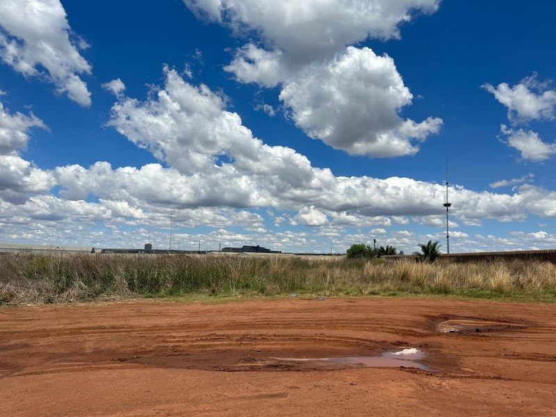 An expansive, unpaved industrial plot featuring red earth, natural vegetation, and distant industrial structures under a clear blue sky.