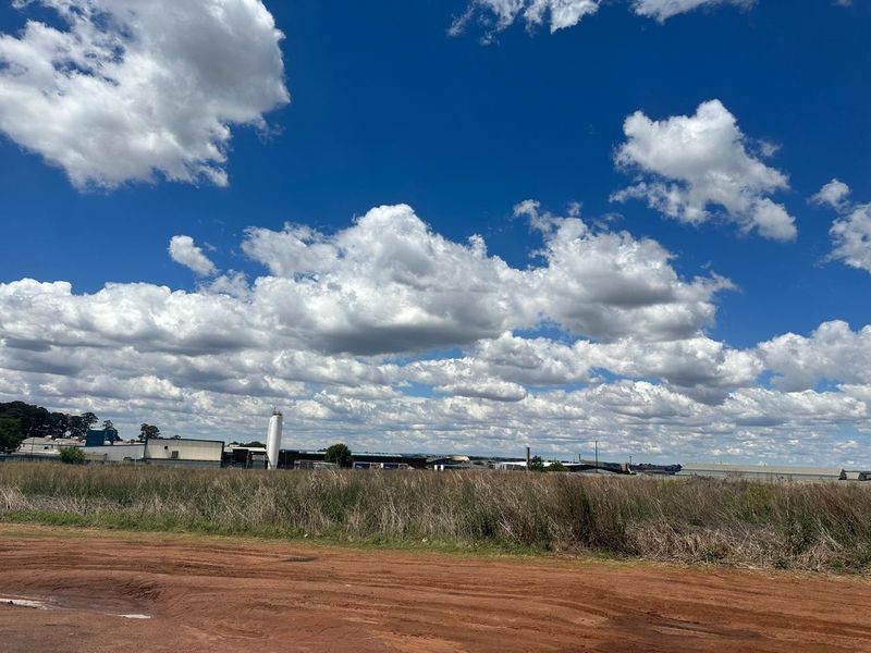 A wide-angle view of an industrial site featuring large warehouse structures, a silo, and surrounding open land under a clear blue sky.