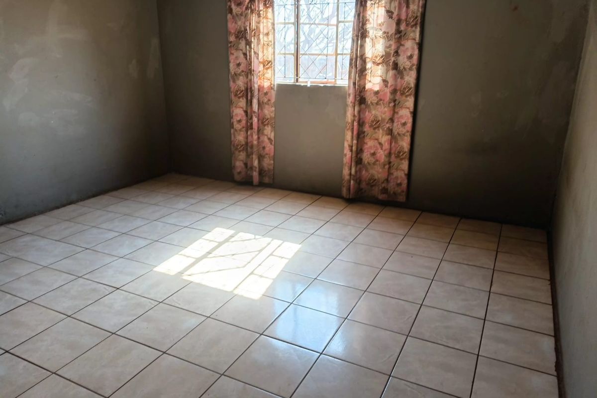 An empty bedroom featuring ceramic tiled flooring and a window with security bars, requiring wall finishing and painting to complete the space.