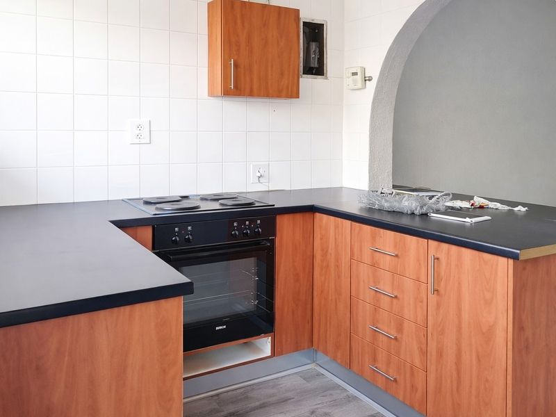 A compact kitchen featuring wood-look cabinetry, dark laminate countertops, a built-in oven, and a hob, finished with white wall tiles and an arched opening.