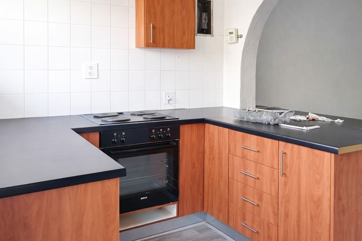 A compact kitchen featuring wood-look cabinetry, dark laminate countertops, a built-in oven, and a hob, finished with white wall tiles and an arched opening.